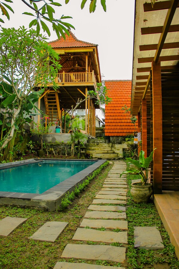 A pool area with a stone pathway leading to a wooden structure with a red roof. - Dedeane Ubud (Ubud)