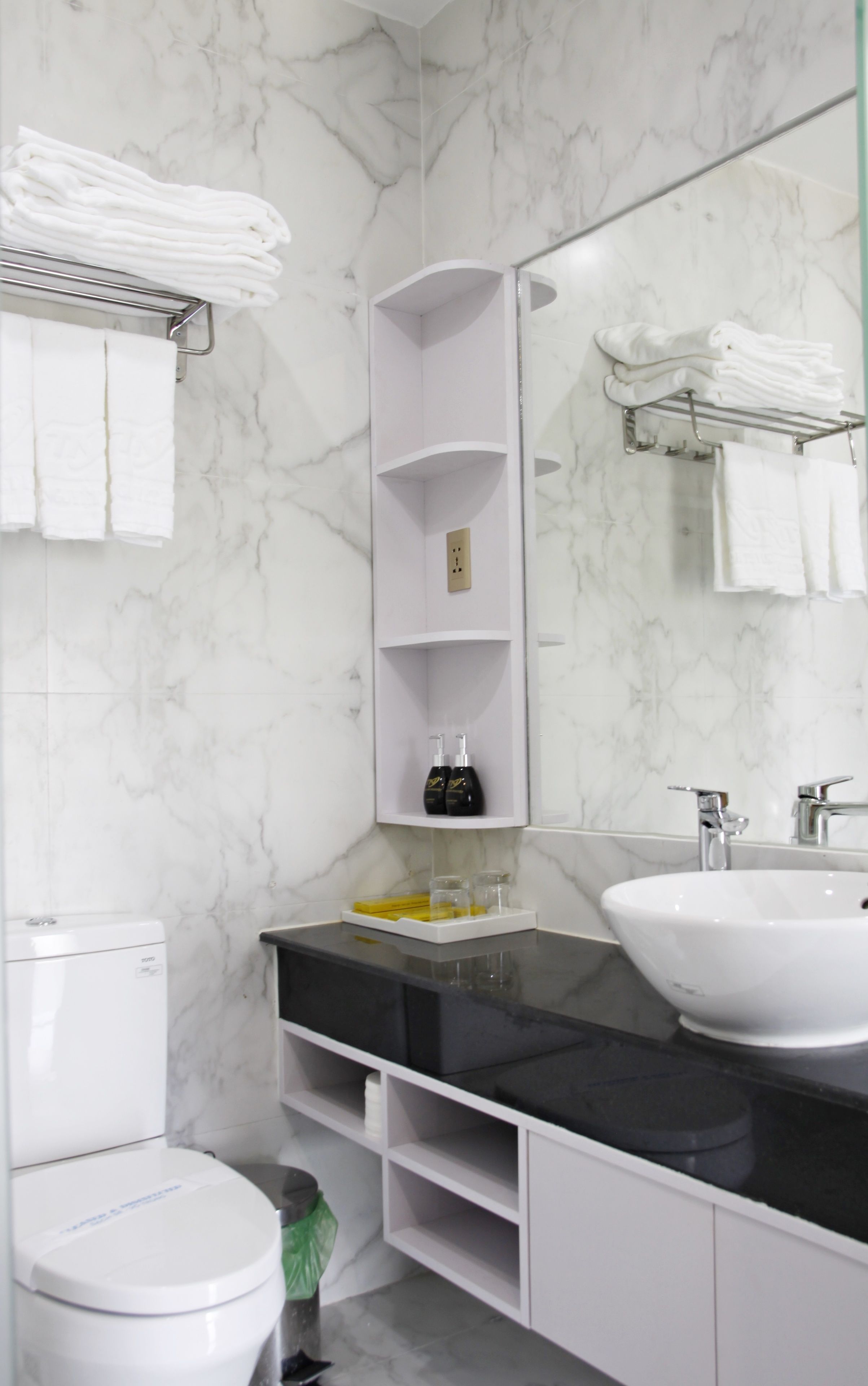 A modern bathroom with a white toilet, a black sink, and a marble backsplash. There are shelves with toiletries and a towel rack above the sink.