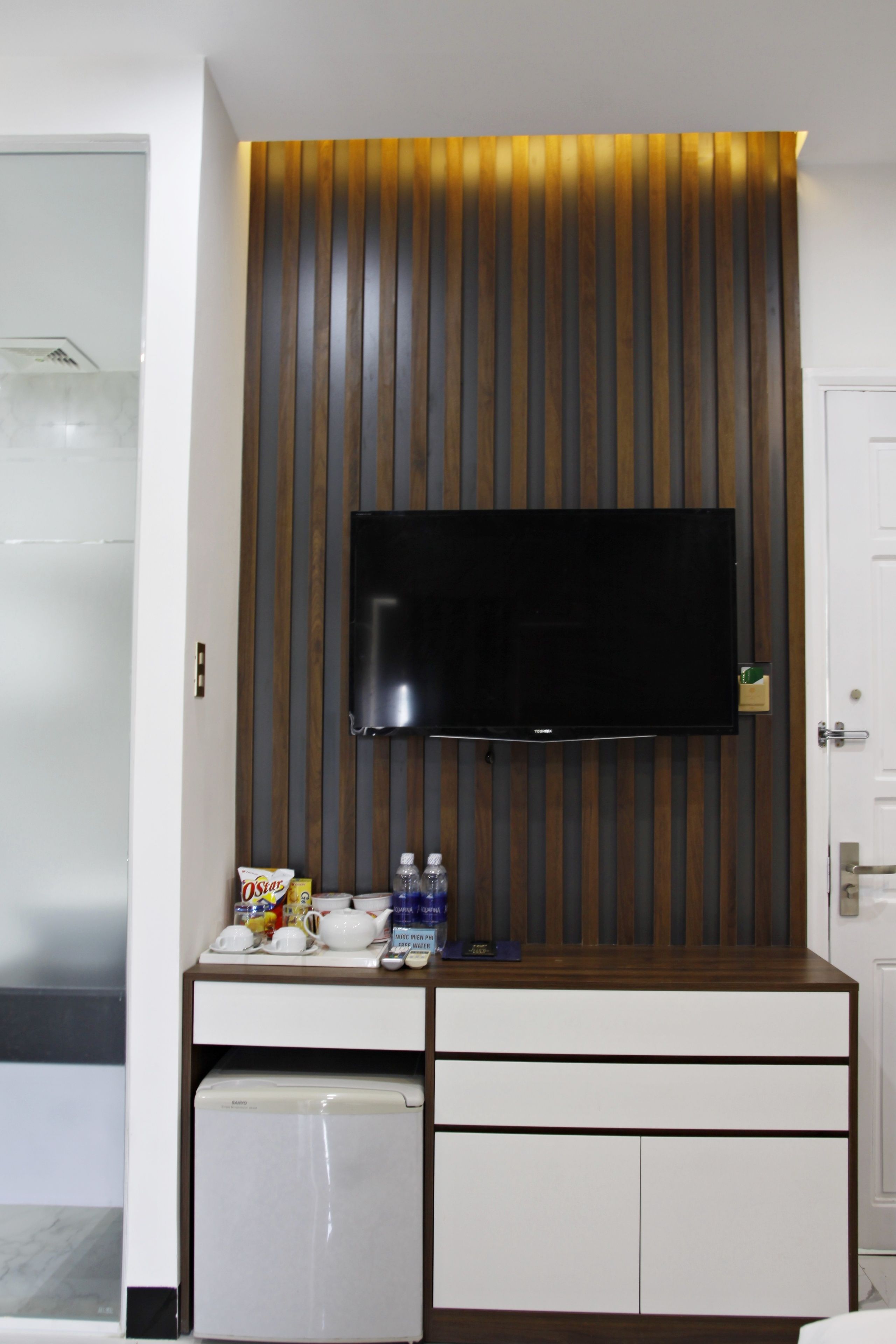 A hotel room with a flat-screen TV mounted on a wooden panel wall, a minibar, and a small refrigerator.