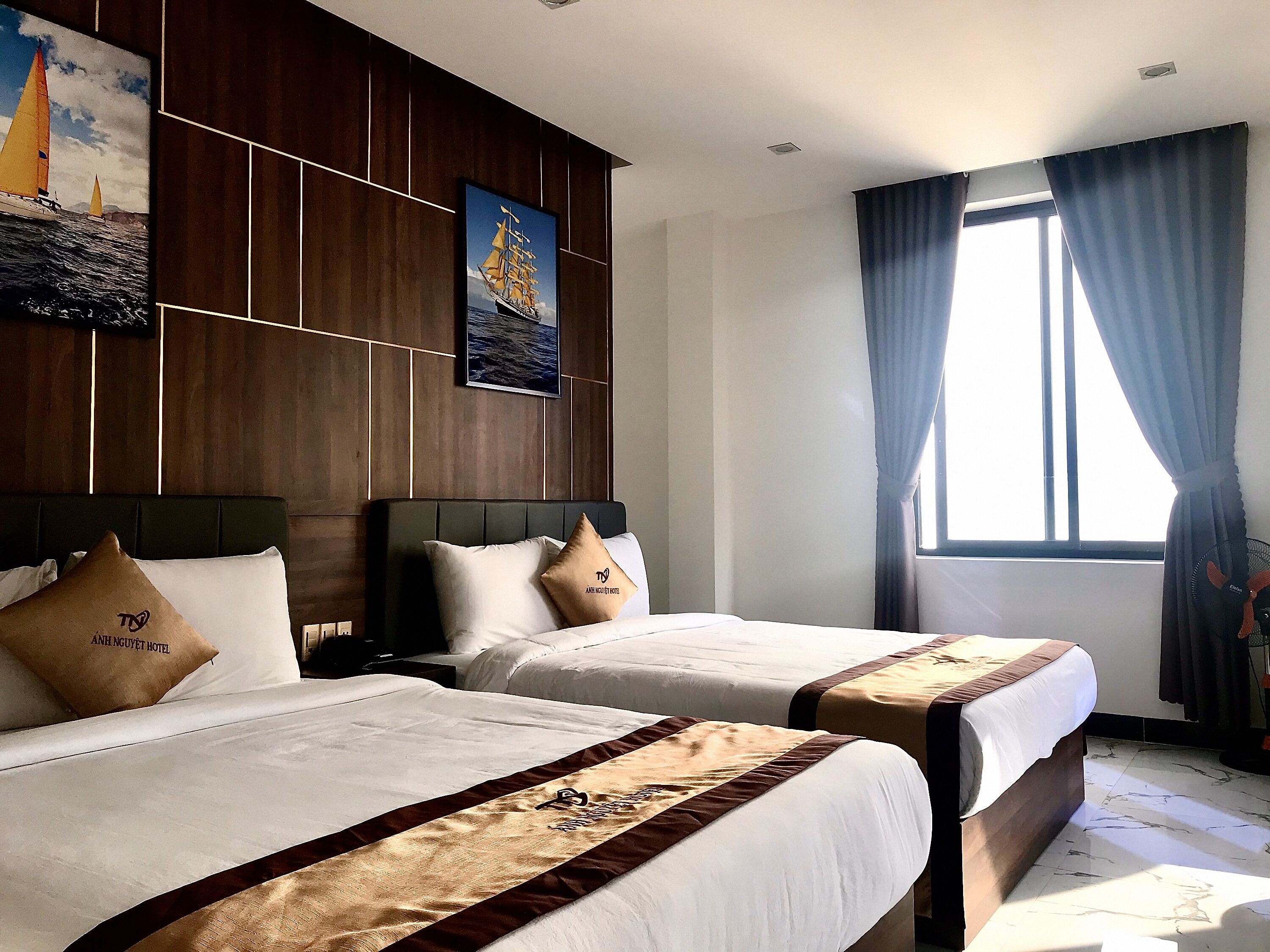 A hotel room with two beds, wooden paneling on the walls, a large window with curtains, and artwork depicting sailing ships.