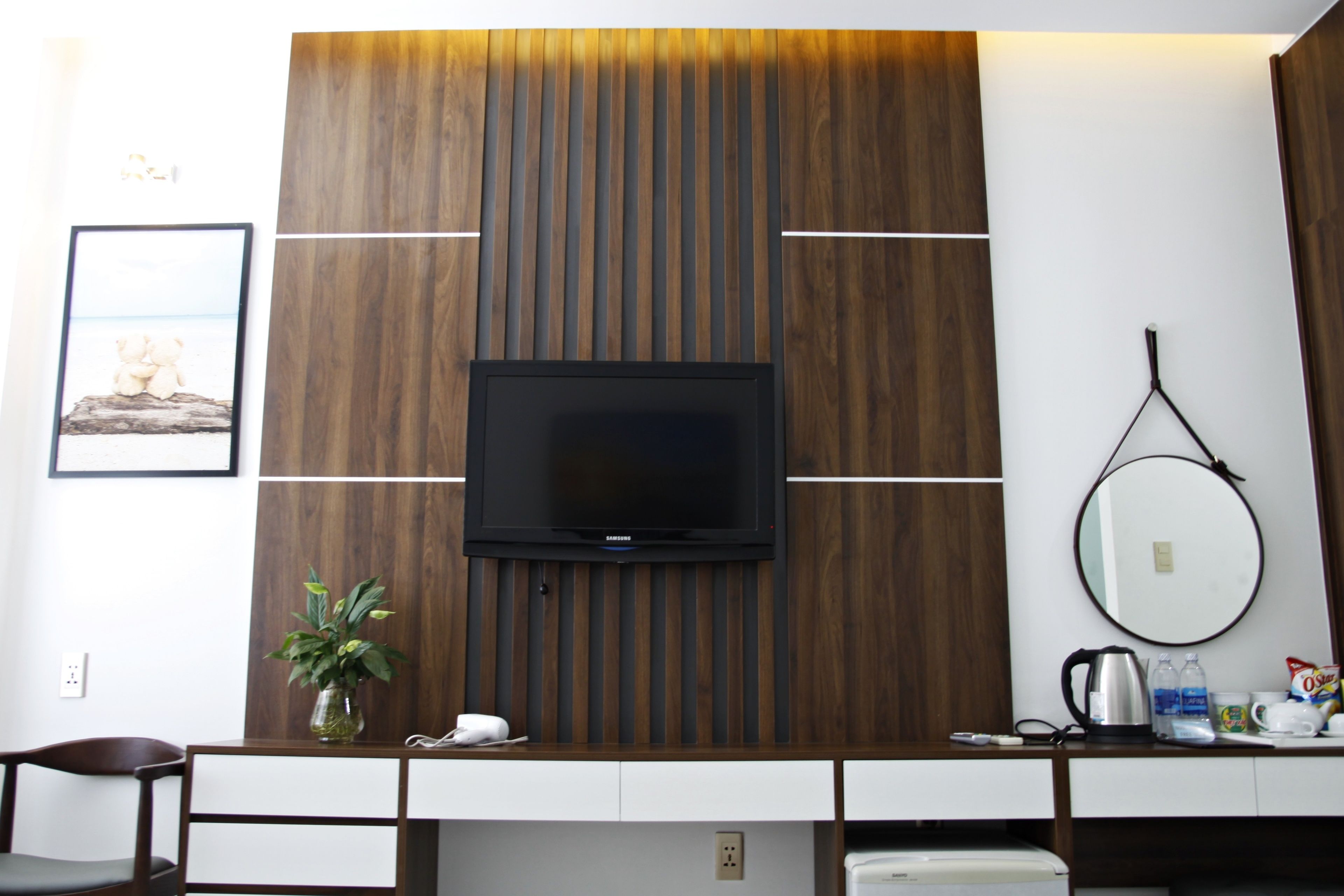 Habitación de hotel con una televisión de pantalla plana montada en una pared de paneles de madera, una mesita con un florero, una cafetera y un espejo en la pared.