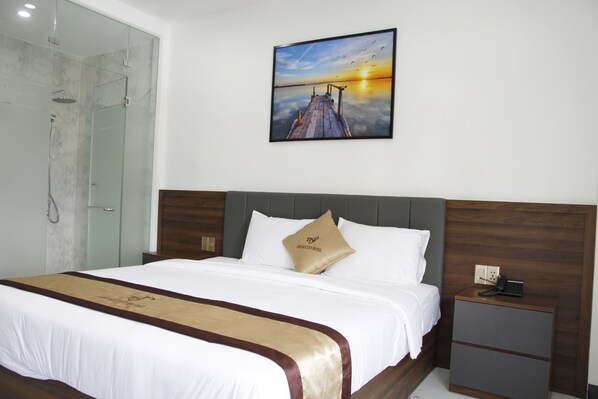A hotel room with a bed, a nightstand, a glass shower enclosure, and a painting of a sunset over a pier.