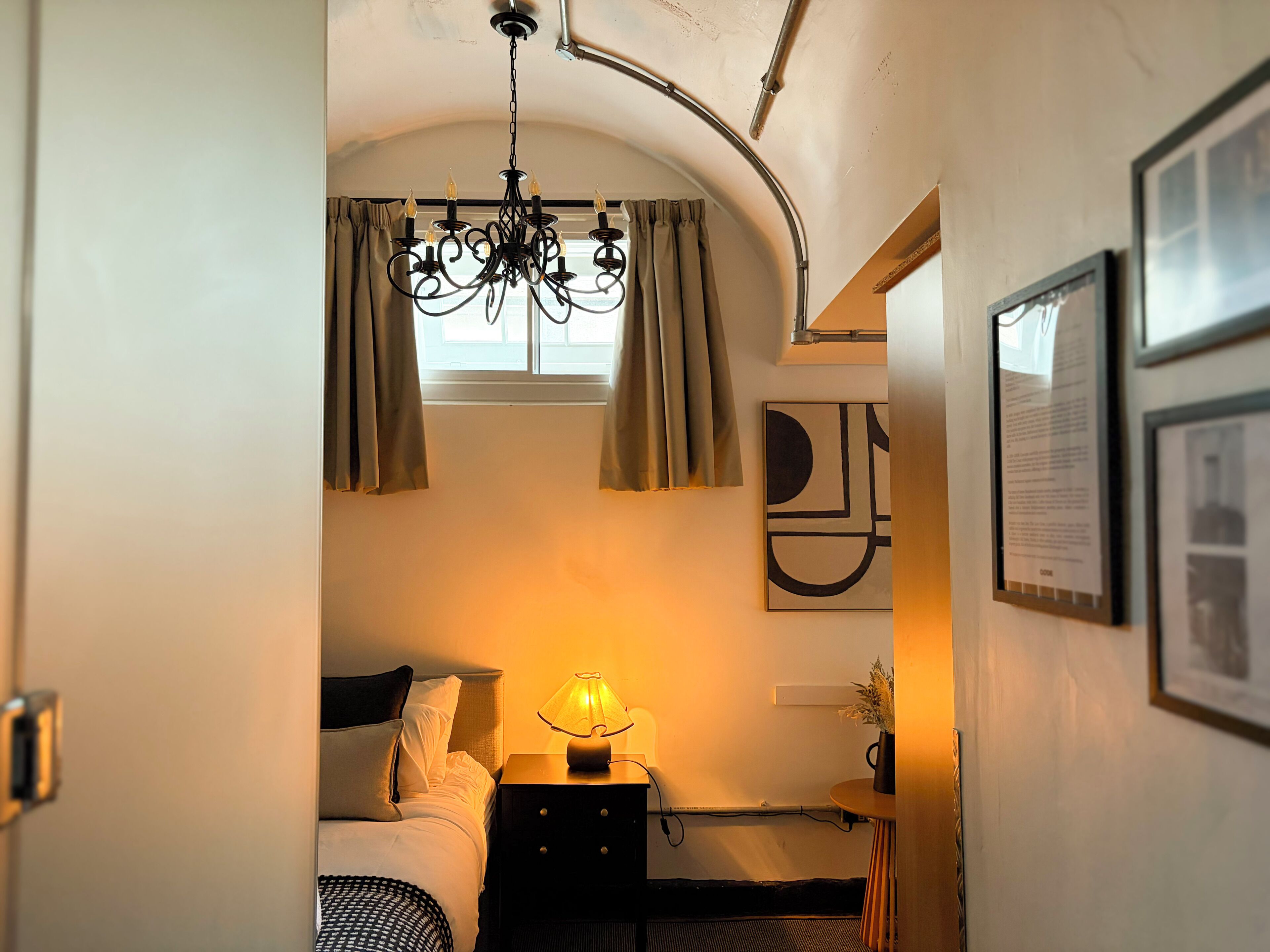 Une chambre à coucher avec un lit, une table de chevet avec une lampe, une fenêtre avec des rideaux, un lustre et une œuvre d’art abstraite accrochée au mur.