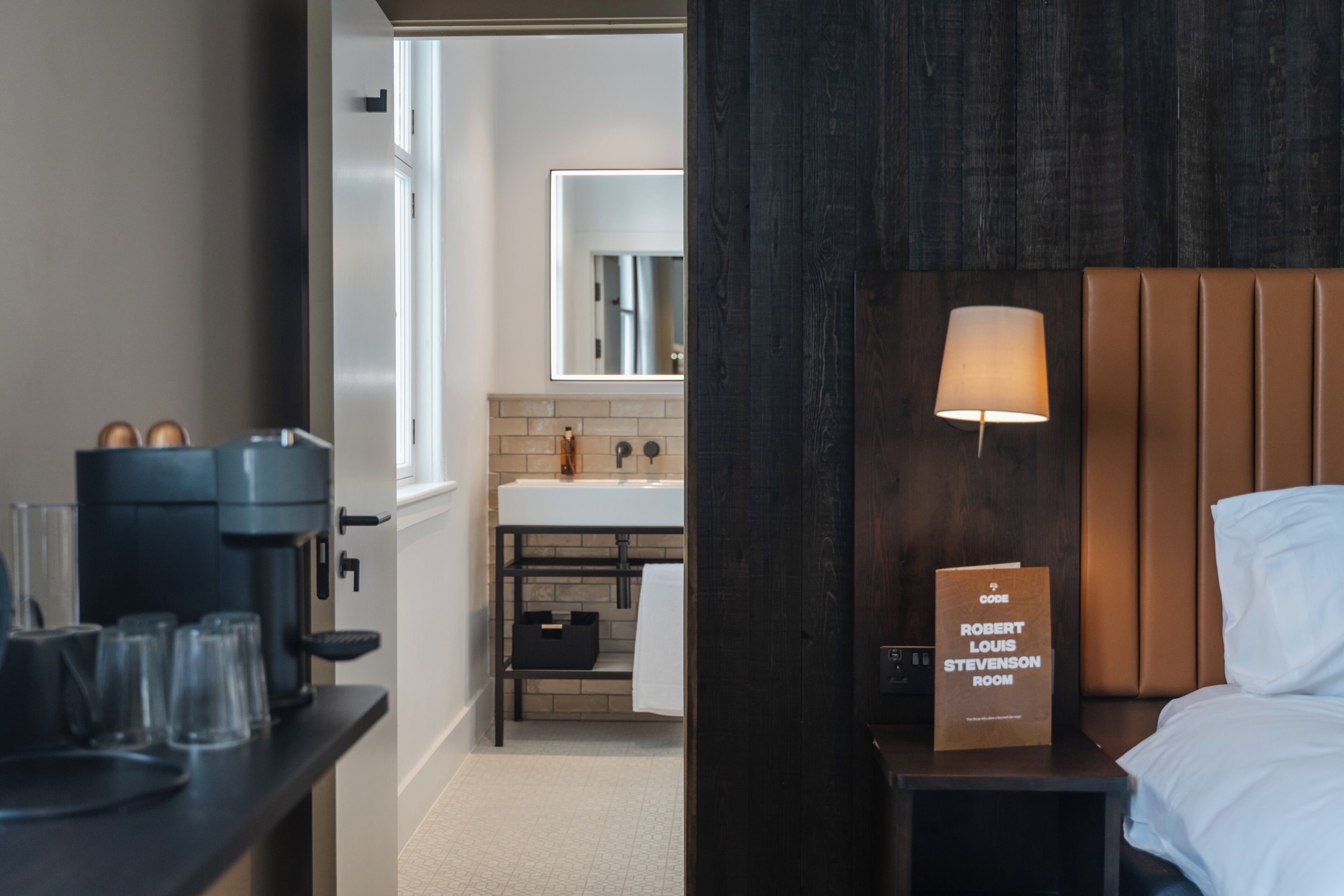 A hotel room with a bed, a bedside table with a lamp, a coffee machine, and a view of the bathroom.