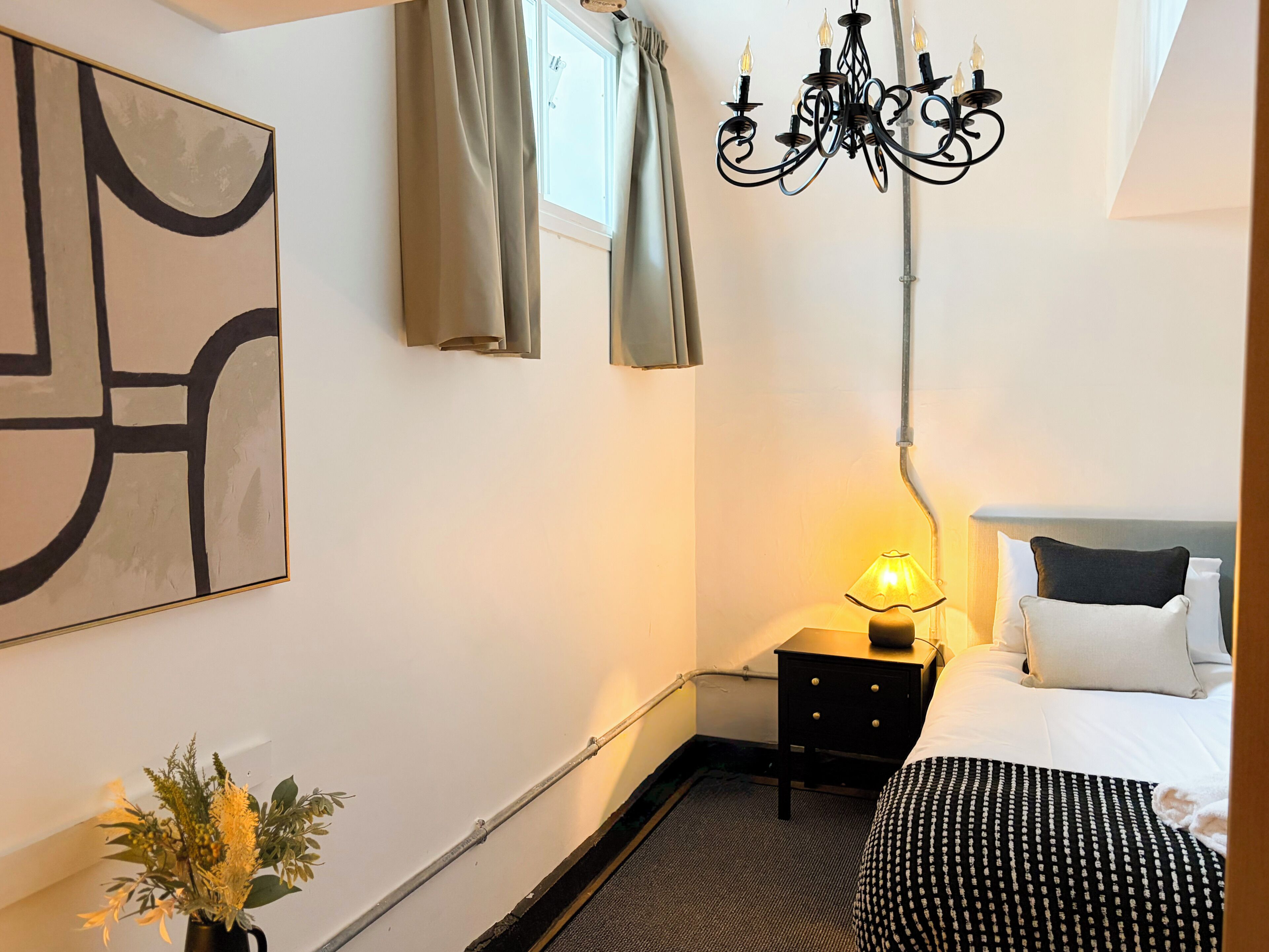 A hotel room with a bed, a nightstand with a lamp, a wall-mounted painting, and a chandelier.