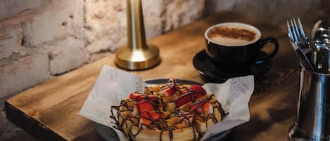 Un plato de gofres con fresas y salsa de chocolate, una taza de café y una lámpara sobre una mesa de madera.