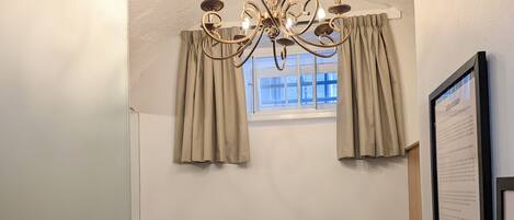 A hotel room with a bed, pillows, a chandelier, curtains, a window, and a welcome sign.