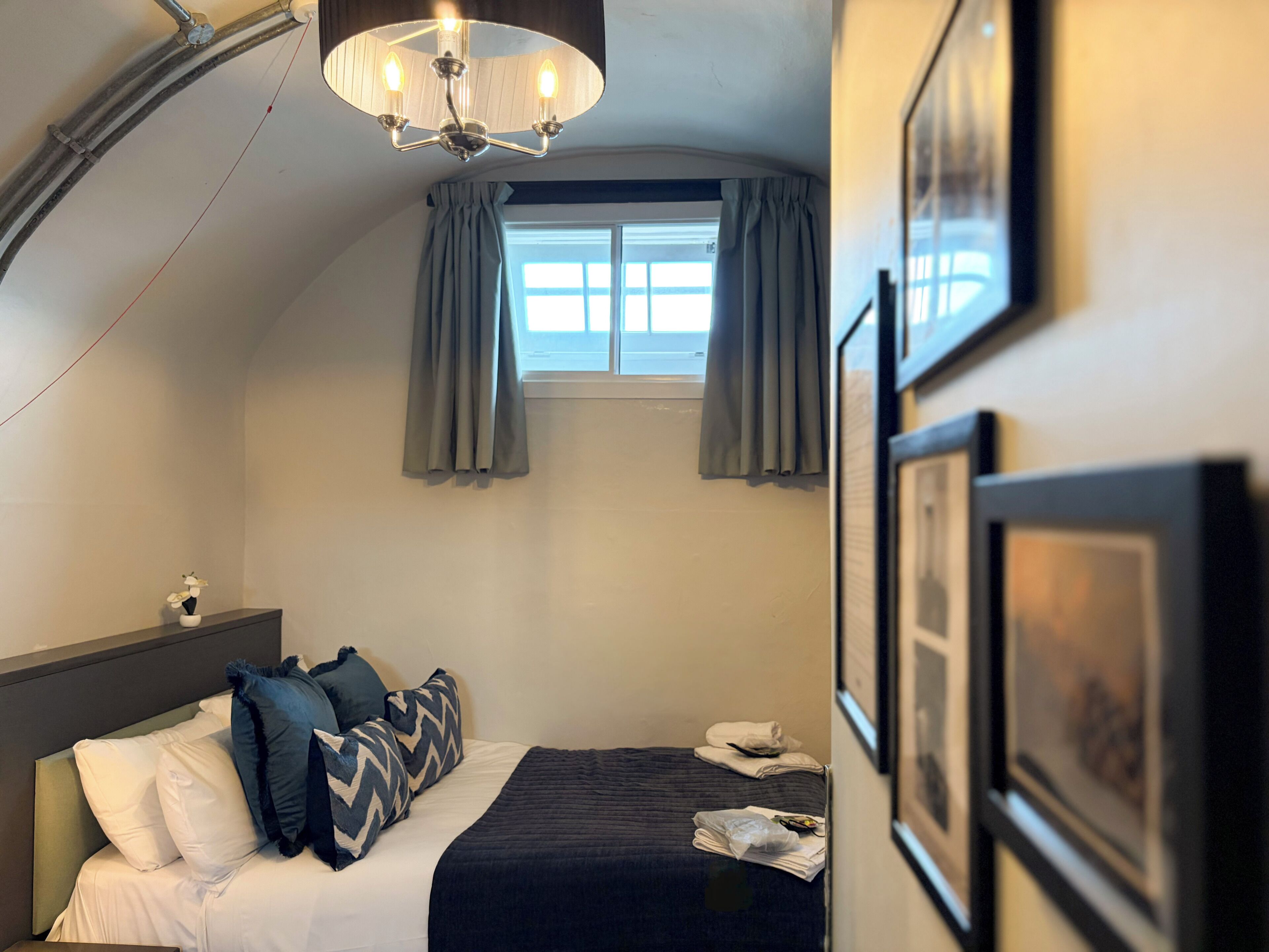 A hotel room with a bed, a nightstand, a window with curtains, and a chandelier.