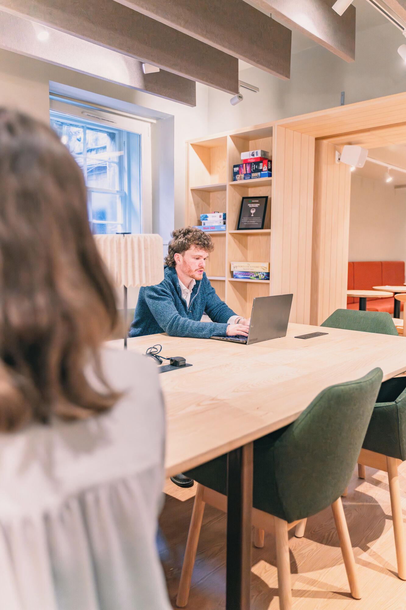 Une personne travaille sur un ordinateur portable, assise à une table en bois dans un bureau moderne, avec des étagères et une fenêtre.