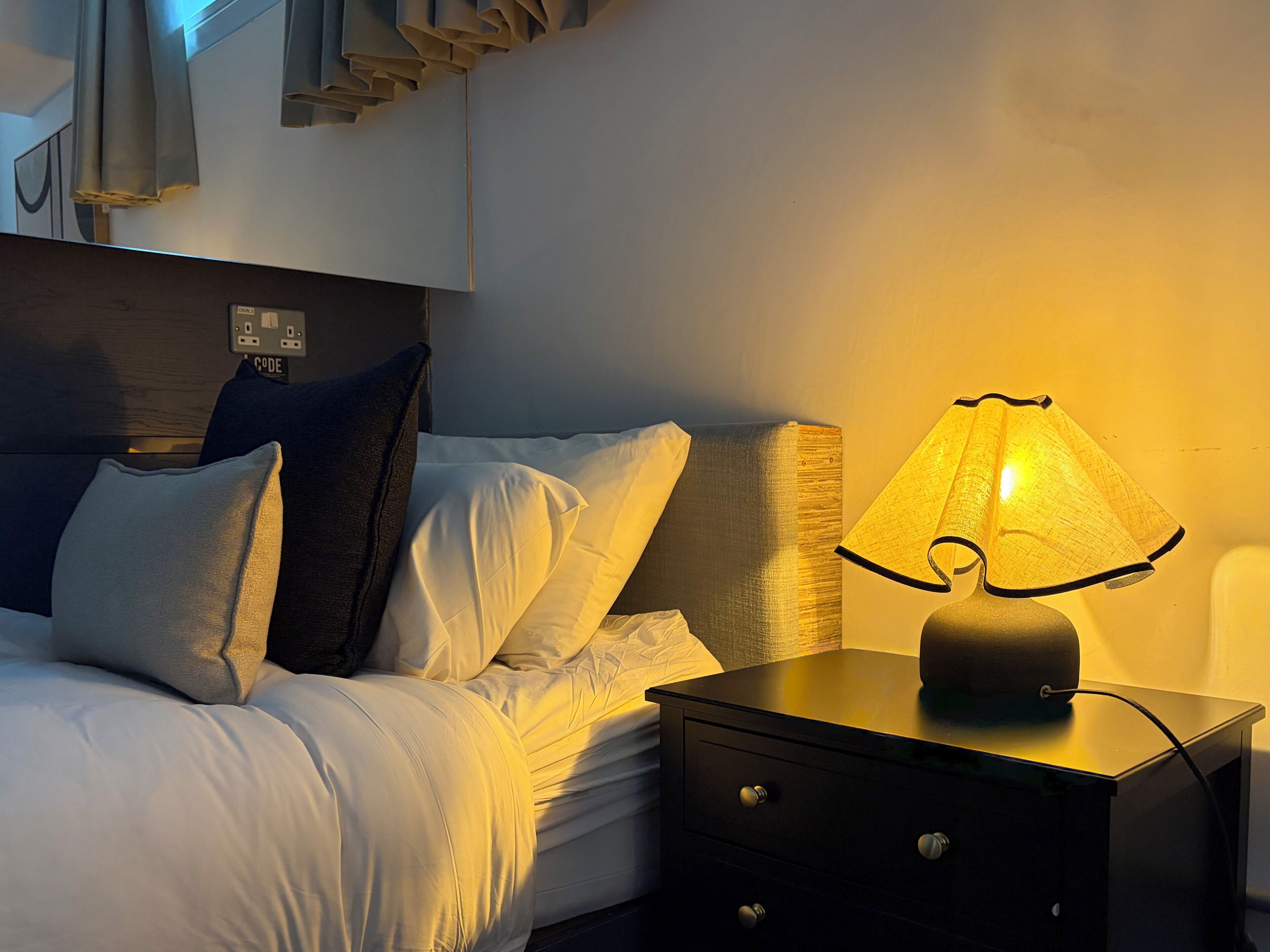 Una cama bien hecha con ropa de cama blanca y varios almohadones, una lámpara de noche y una mesita de noche pequeña de color negro con cajones.