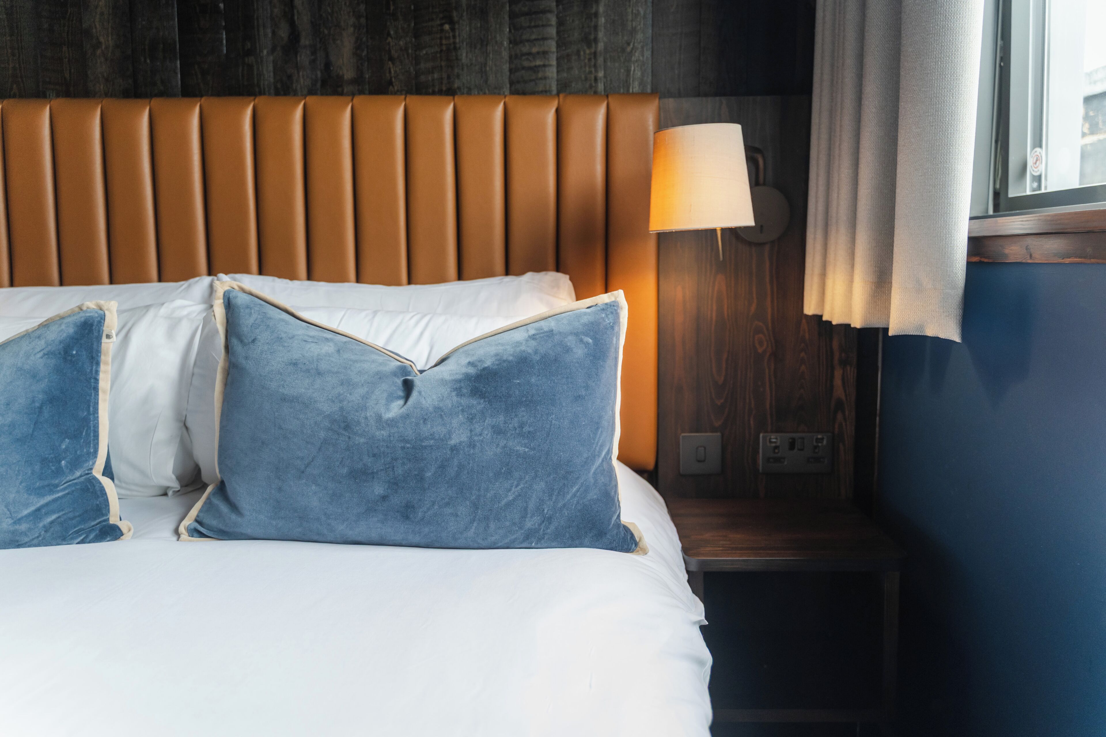 A neatly made bed with white linens and two blue pillows, a wooden headboard, a bedside lamp, and a window with curtains.