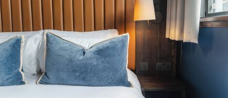 Ein ordentlich bezogenes Bett mit weißen Bettwäsche und zwei blauen Kissen, ein hölzernes Kopfteil, eine Nachttischlampe und ein Fenster mit Vorhängen.