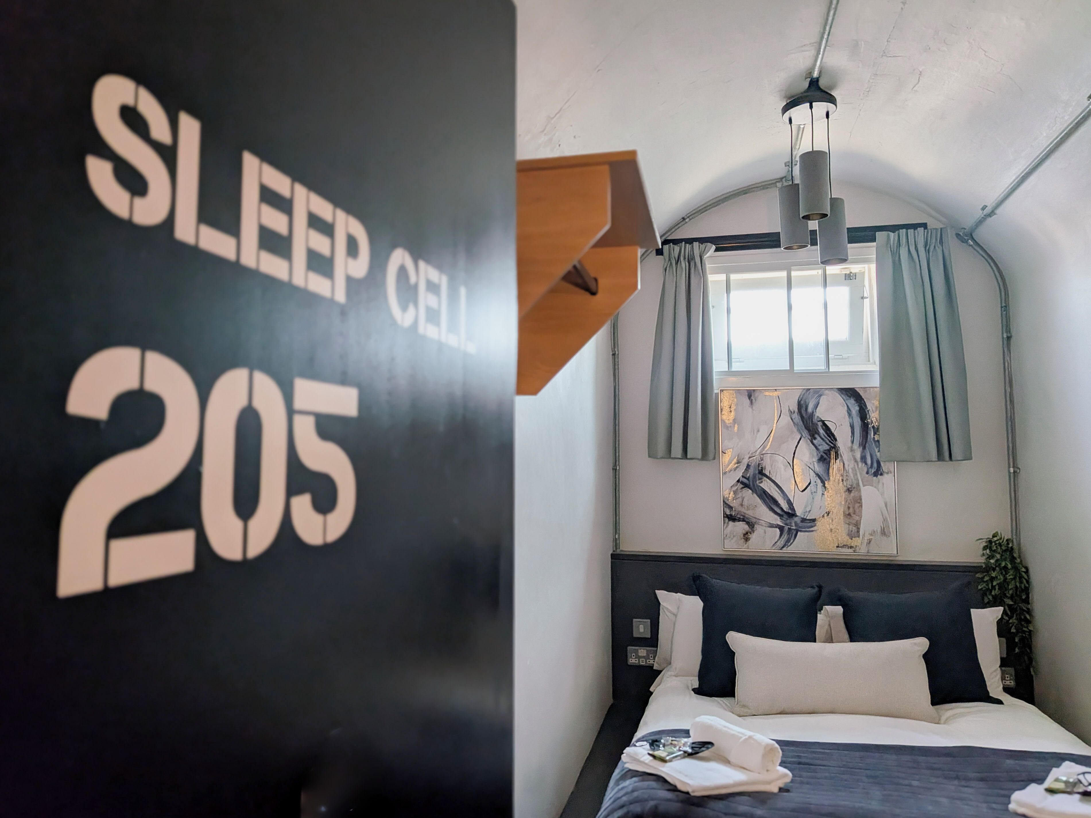 A modern hotel room with a bed, a painting, and a sign indicating 'SLEEP CELL 205'.