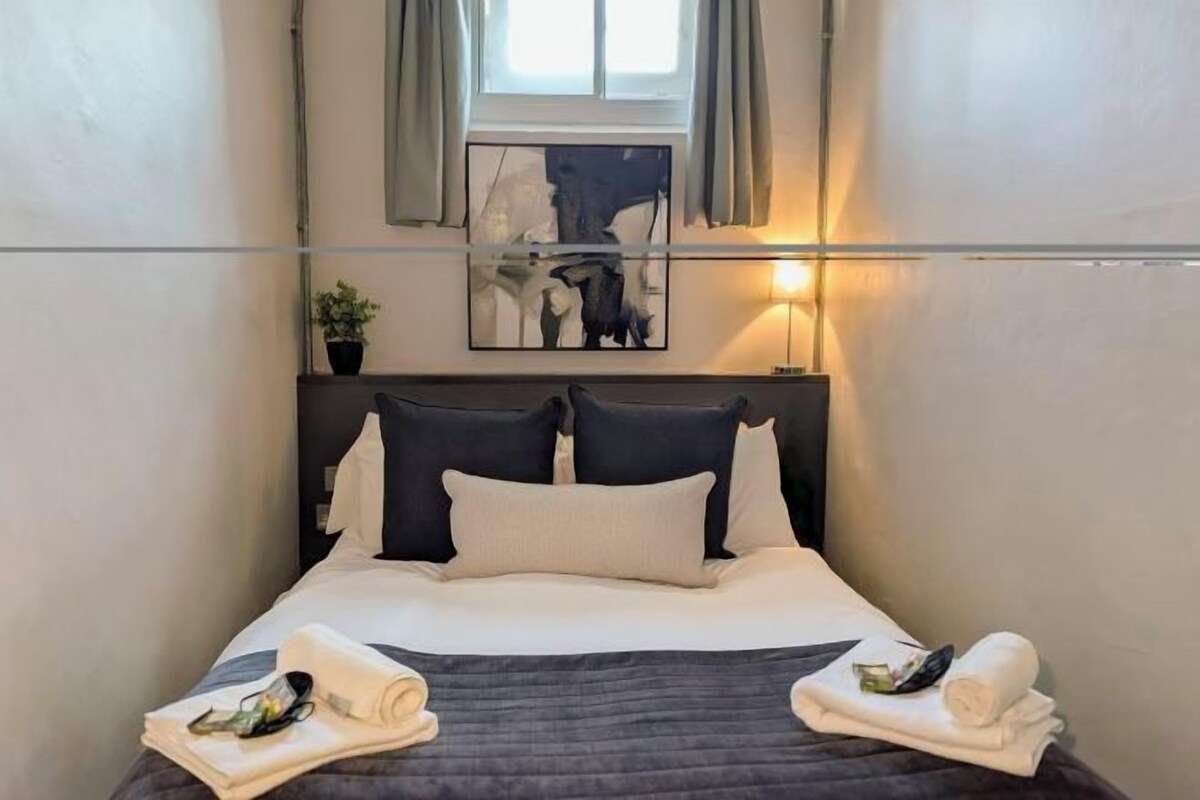 A neatly made bed with pillows and towels, a bedside lamp, a window with curtains, and a wall-mounted artwork.