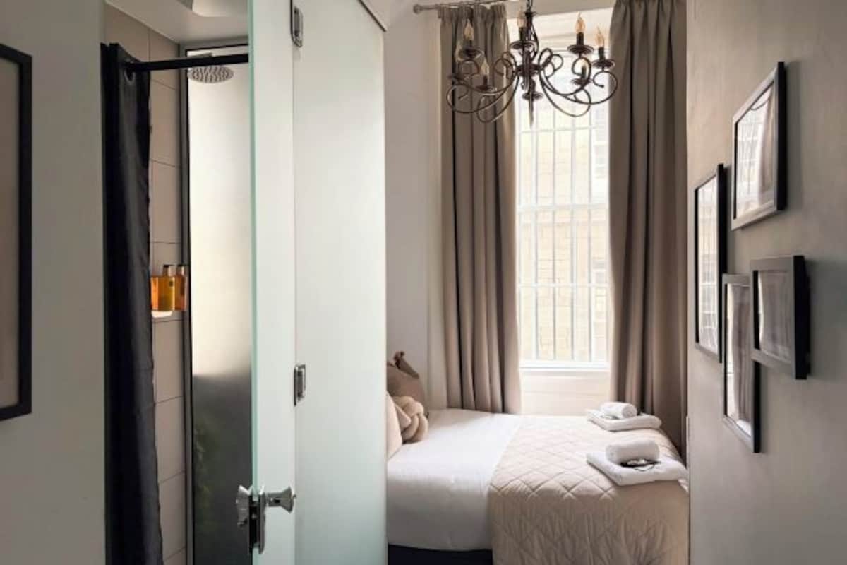 A bedroom with a bed, a window with curtains, a chandelier, and framed pictures on the wall.
