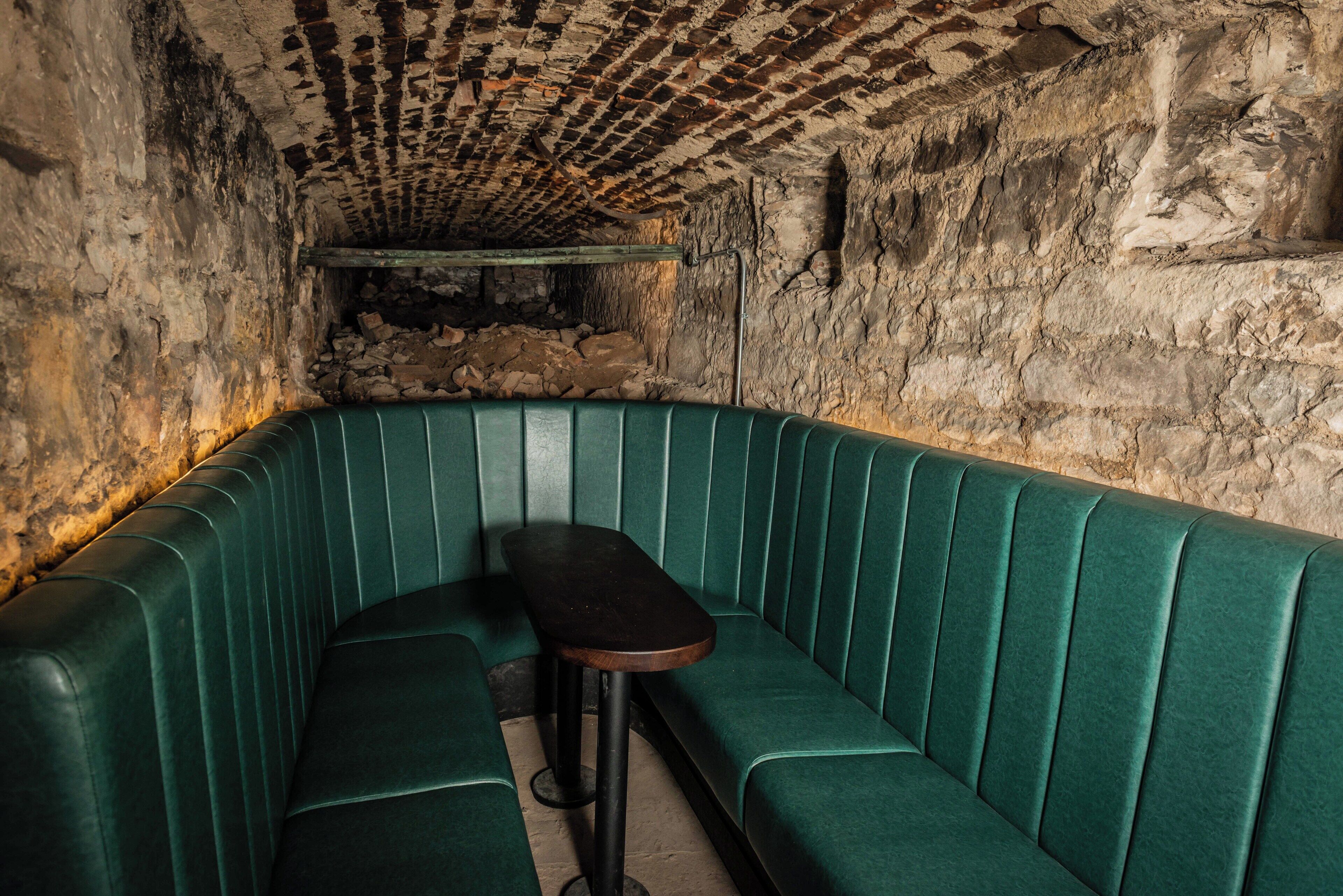A green upholstered booth with a wooden table and bench, set against a textured stone wall.