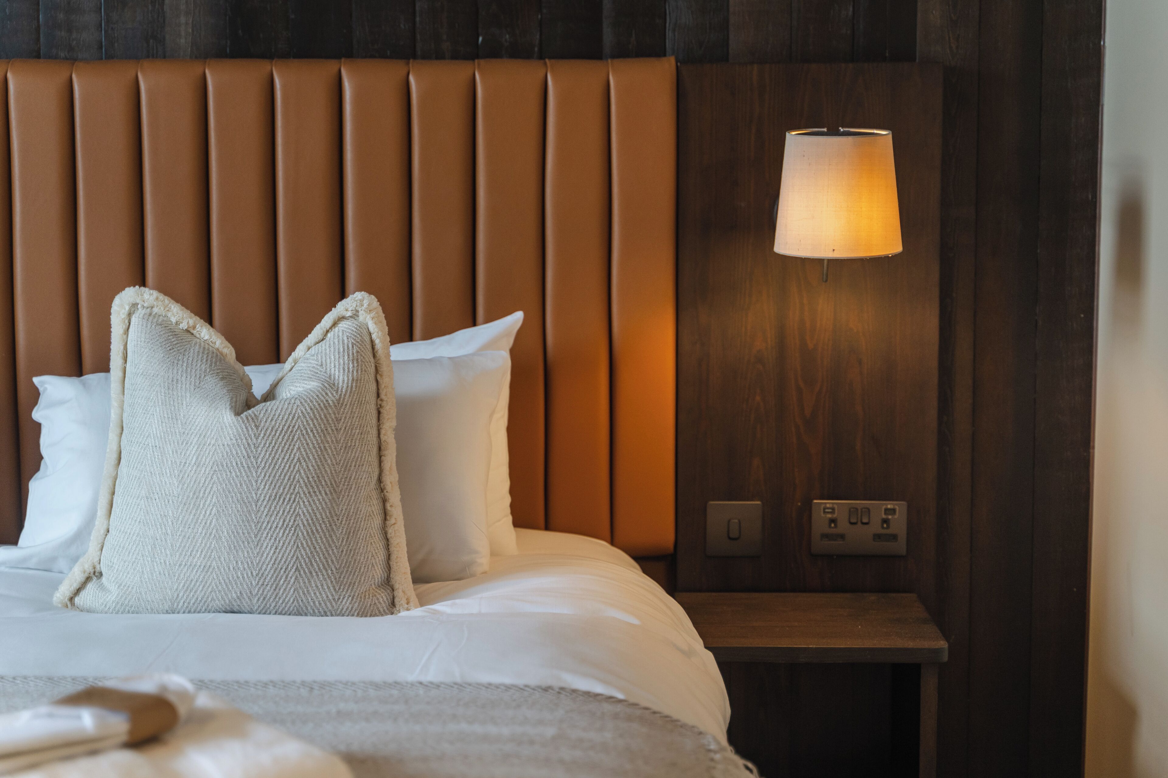 A hotel room with a bed, pillows, a nightstand, a lamp, and a power outlet.