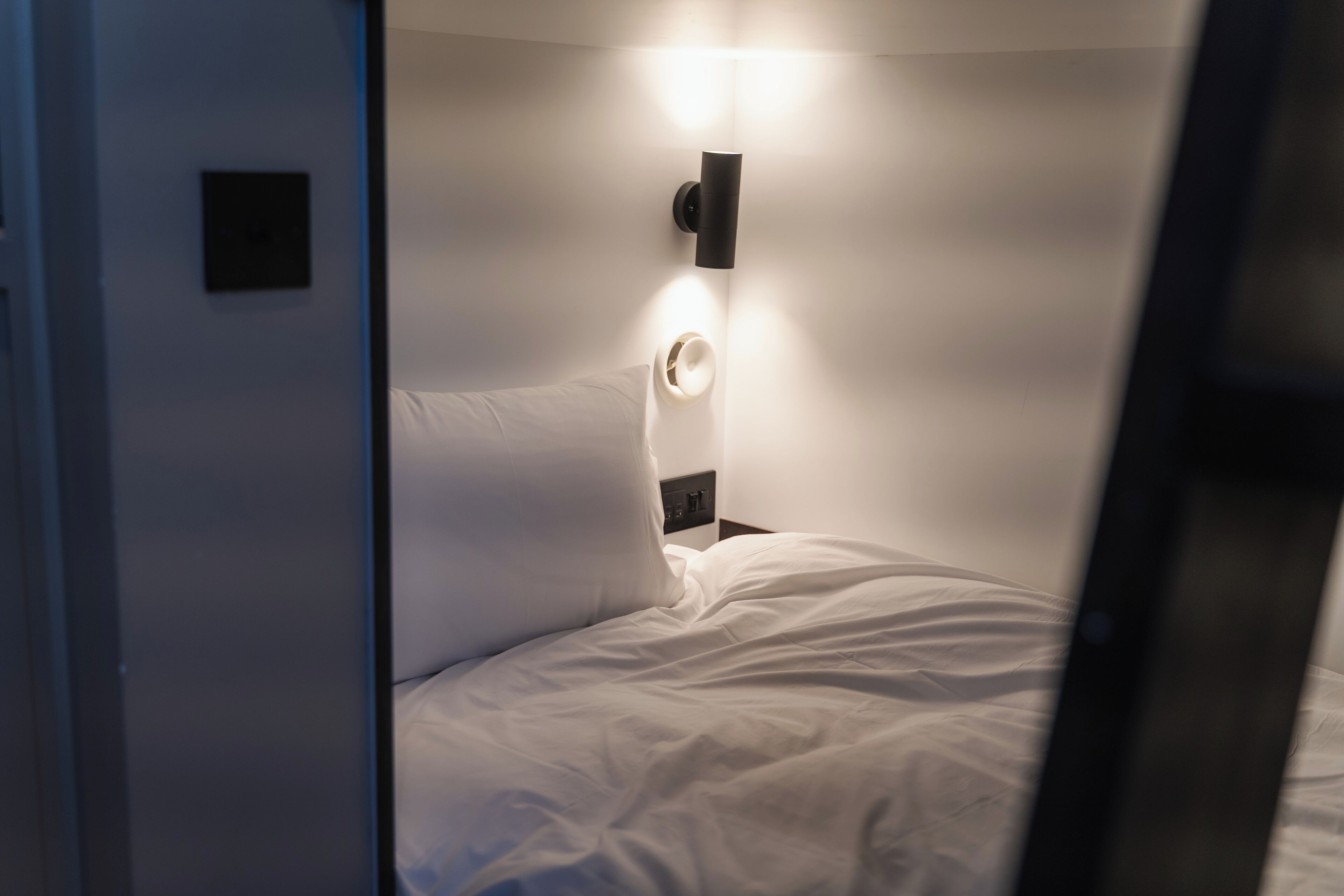 Une petite pièce faiblement éclairée, avec un lit simple, une lampe fixée au mur et une petite étagère sur laquelle se trouve un vase.