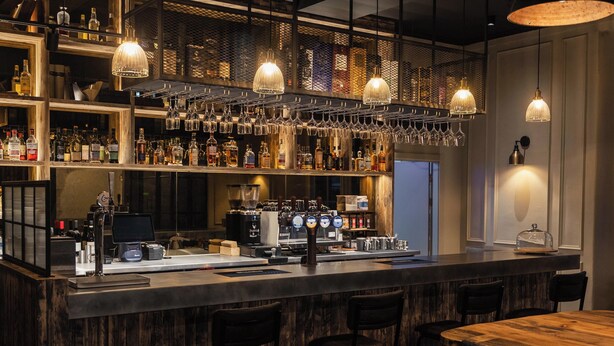 A bar area with wooden shelves stocked with bottles, a bar counter with a coffee machine, and hanging light fixtures. - CODE - The Court, Edinburgh (Edinburgh)