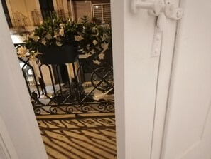 Una habitación con piso estampado, una silla de metal negro, una planta en maceta con flores blancas y una puerta blanca con manija.