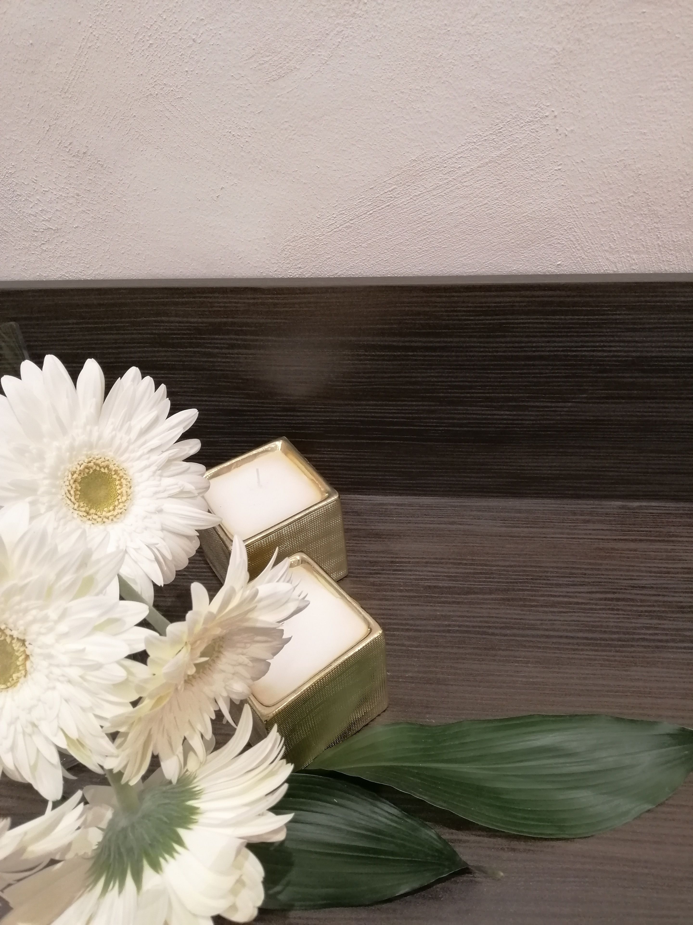 Superficie de madera oscura con dos velas cuadradas en candelabros dorados y flores blancas con hojas verdes.