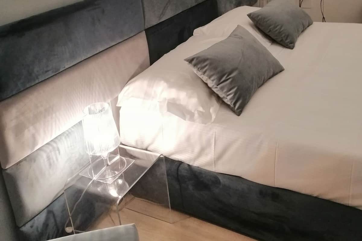 Una cama bien tendida con un cabecero gris y blanco, una lámpara de noche y dos almohadas.