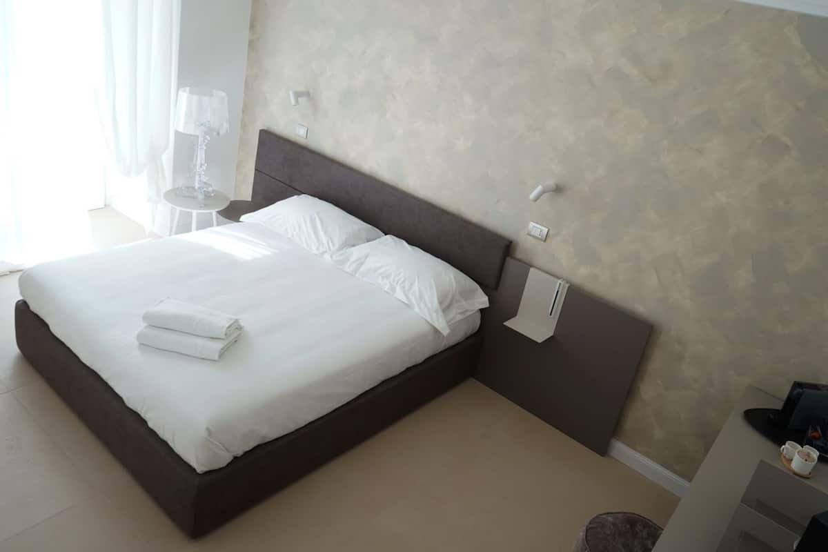 Una cama bien hecha con sábanas blancas y una toalla doblada, una mesita de noche con lámpara, y un termostato fijado a la pared.