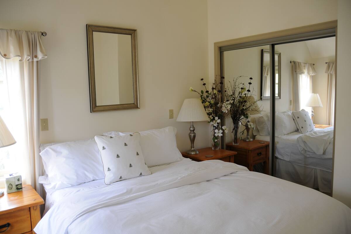 A neatly made bed with white linens and decorative pillows, a bedside table with a lamp and a vase of flowers, and a large mirror reflecting the room.