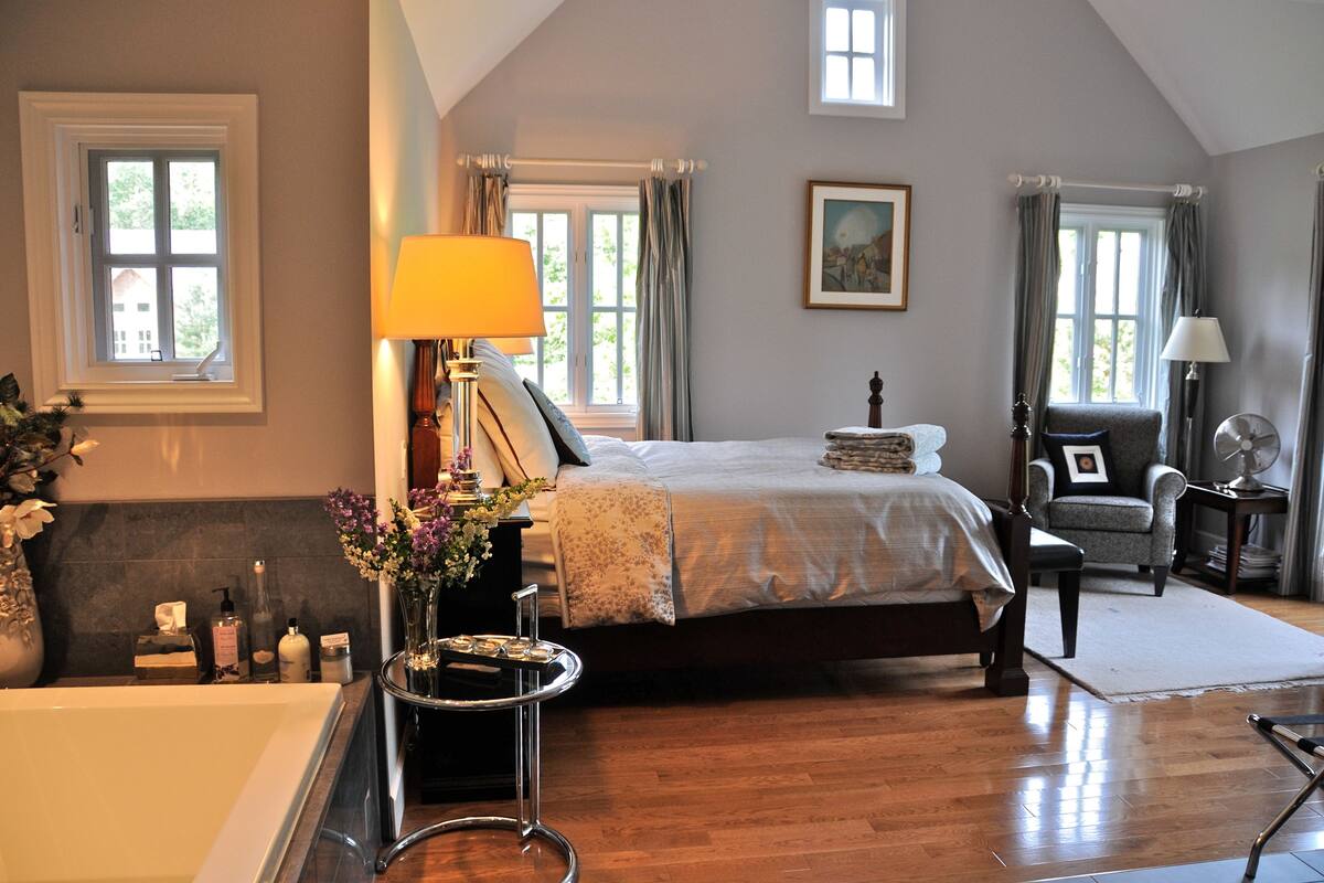 A bedroom with a large bed, a sitting area with a chair, a side table, a lamp, and a window.