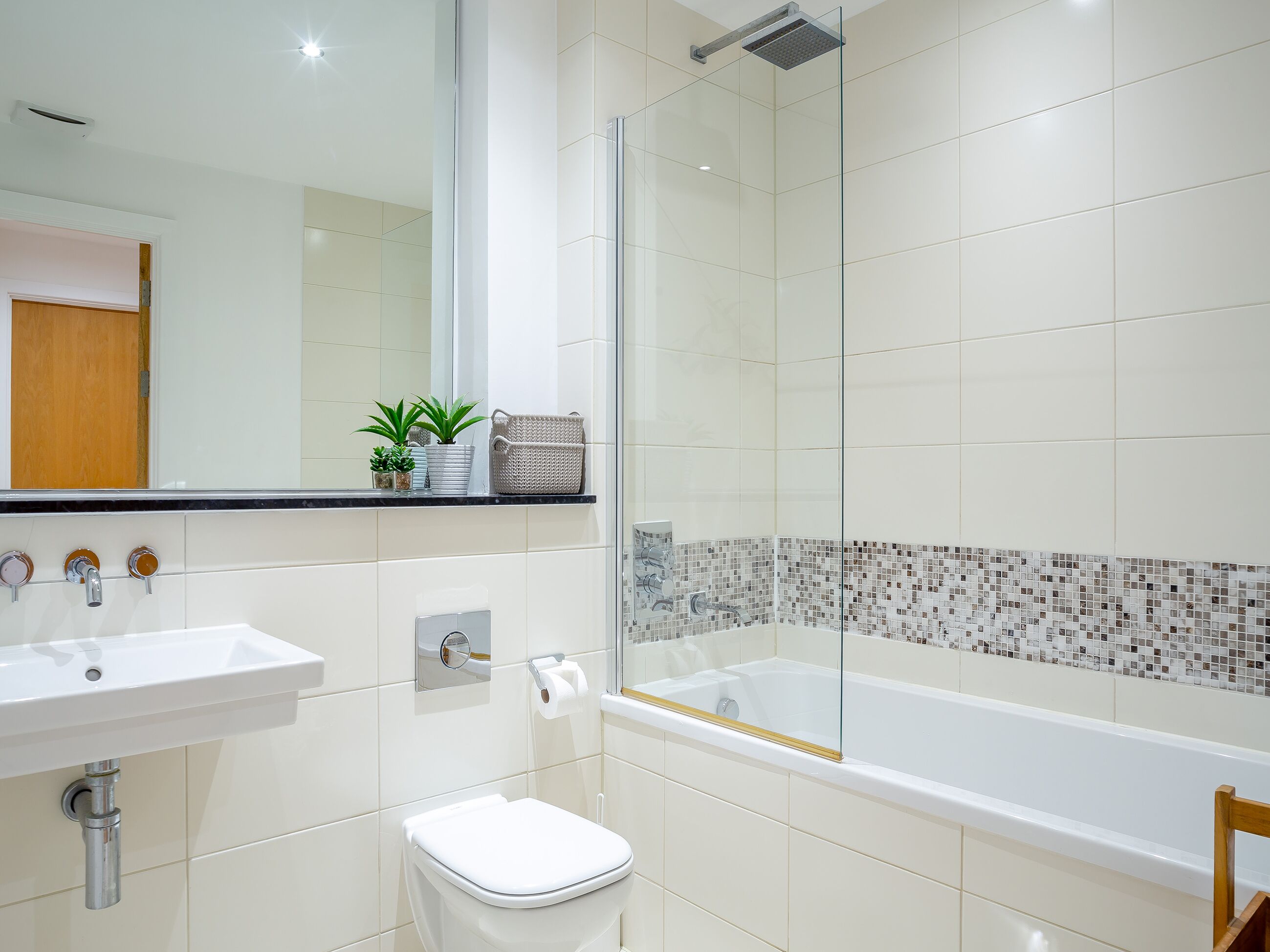 Une salle de bain moderne avec un lavabo, des toilettes et une baignoire blancs. Il y a un espace douche avec une porte en verre et un mur recouvert de carreaux.