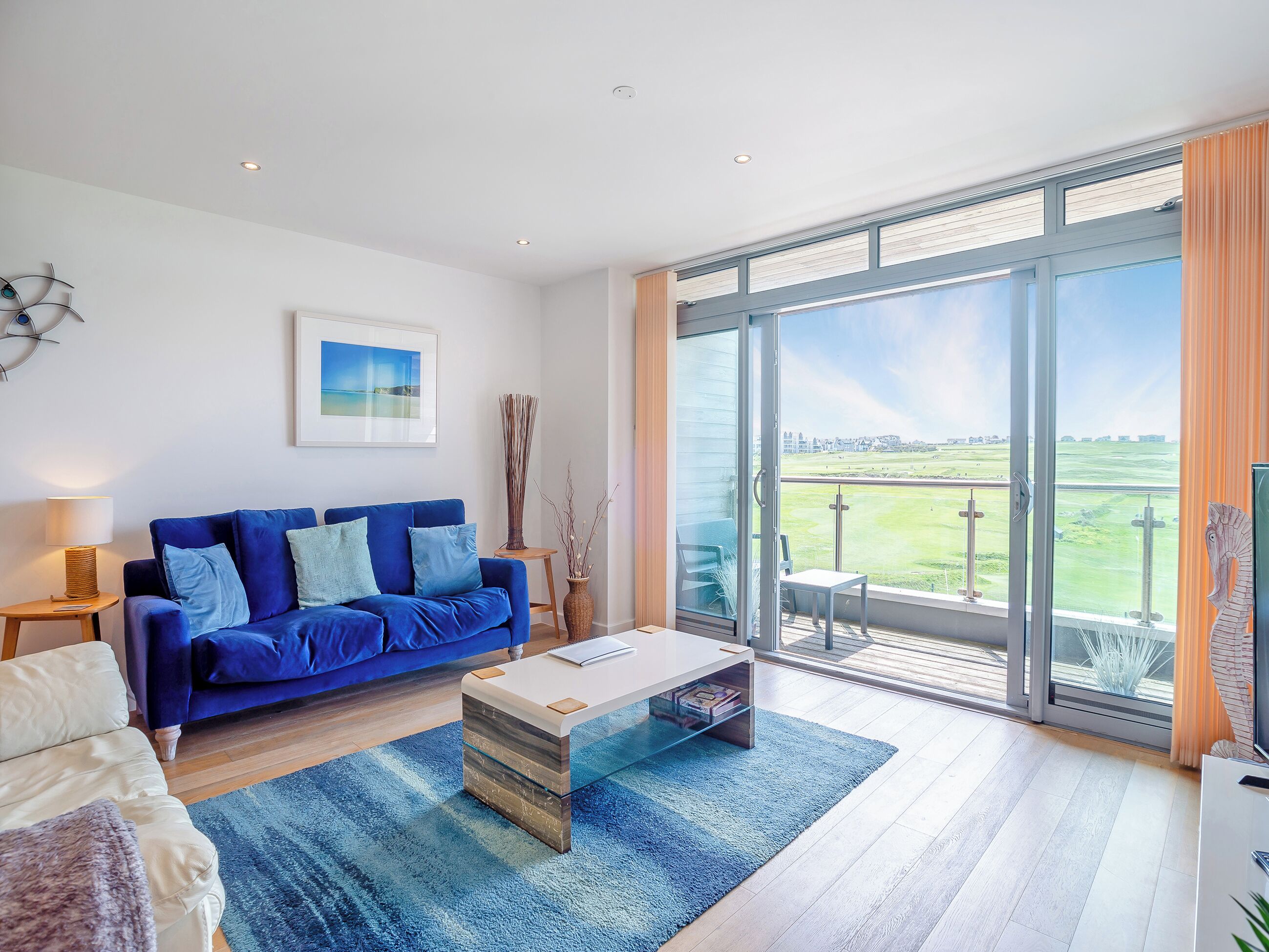 A modern living room with a blue sofa, a glass coffee table, and a view of a golf course through large sliding glass doors.