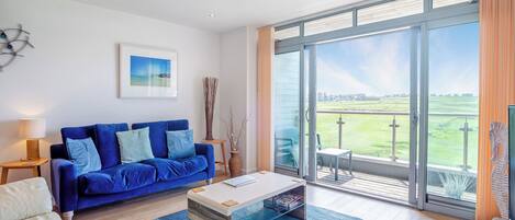 A modern living room with a blue sofa, a glass coffee table, and a view of a golf course through large sliding glass doors.