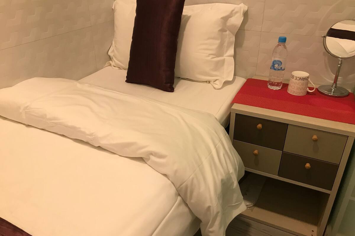 A hotel room with a bed, a nightstand with a mirror, a red tray, and a bottle of water.