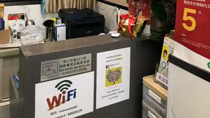A hotel reception desk with a computer, a printer, and a vase of purple orchids. There are signs indicating a 24-hour CCTV system and WiFi availability. - Vve Vve Hotel (Kowloon)