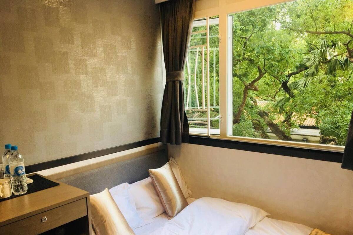 A hotel room with a bed, a nightstand, a window with a view of trees, and a wall with a textured pattern.