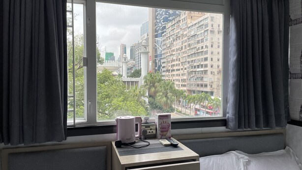 A hotel room with a bed, a desk with a coffee maker, and a view of tall buildings and palm trees outside. - Vve Vve Hotel (Kowloon)