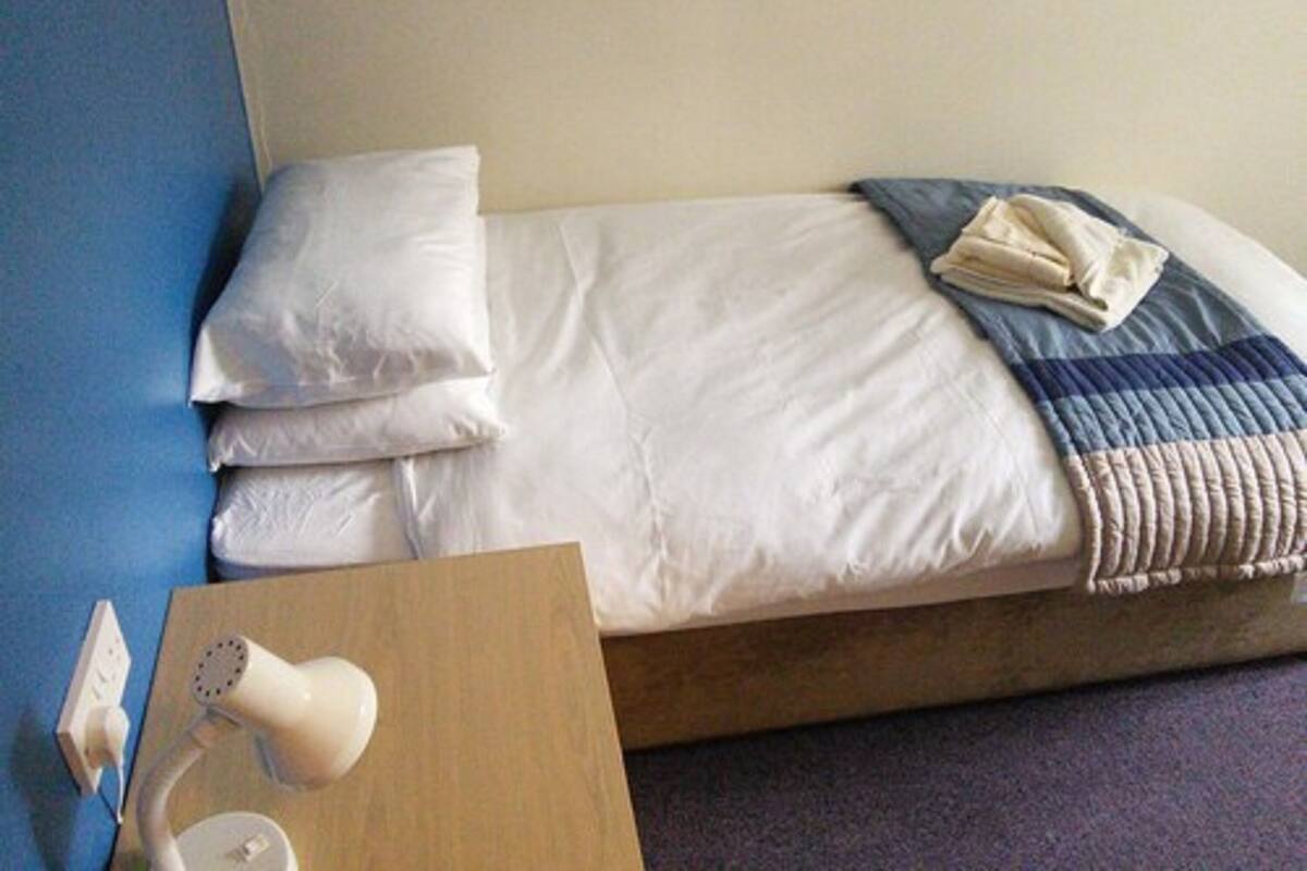 Cama bem arrumada com lençóis brancos e manta listrada em azul e branco, mesa de cabeceira com abajur e parede azul.