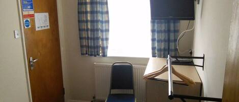 A small, sparsely furnished room with a wooden door, a window with plaid curtains, a radiator, a desk with a computer monitor, and a chair.