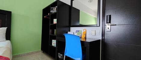 A hotel room with a bed, a desk with a chair, a bookshelf, a mirror, and a door.