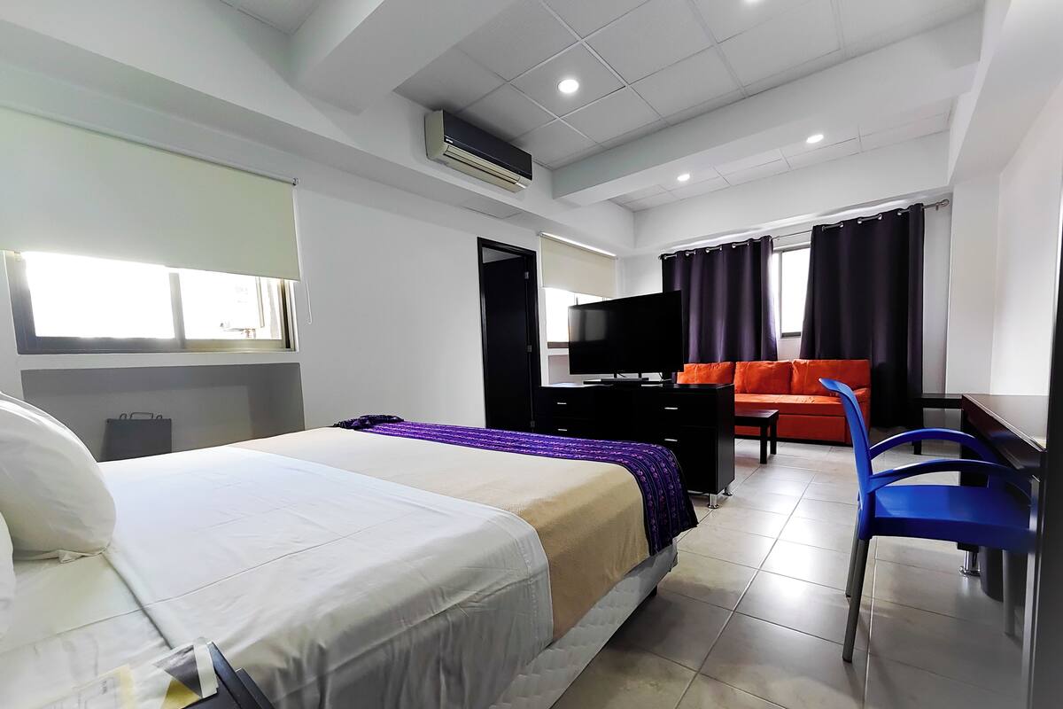 A hotel room with a bed, a desk with a television, a chair, and a sofa.