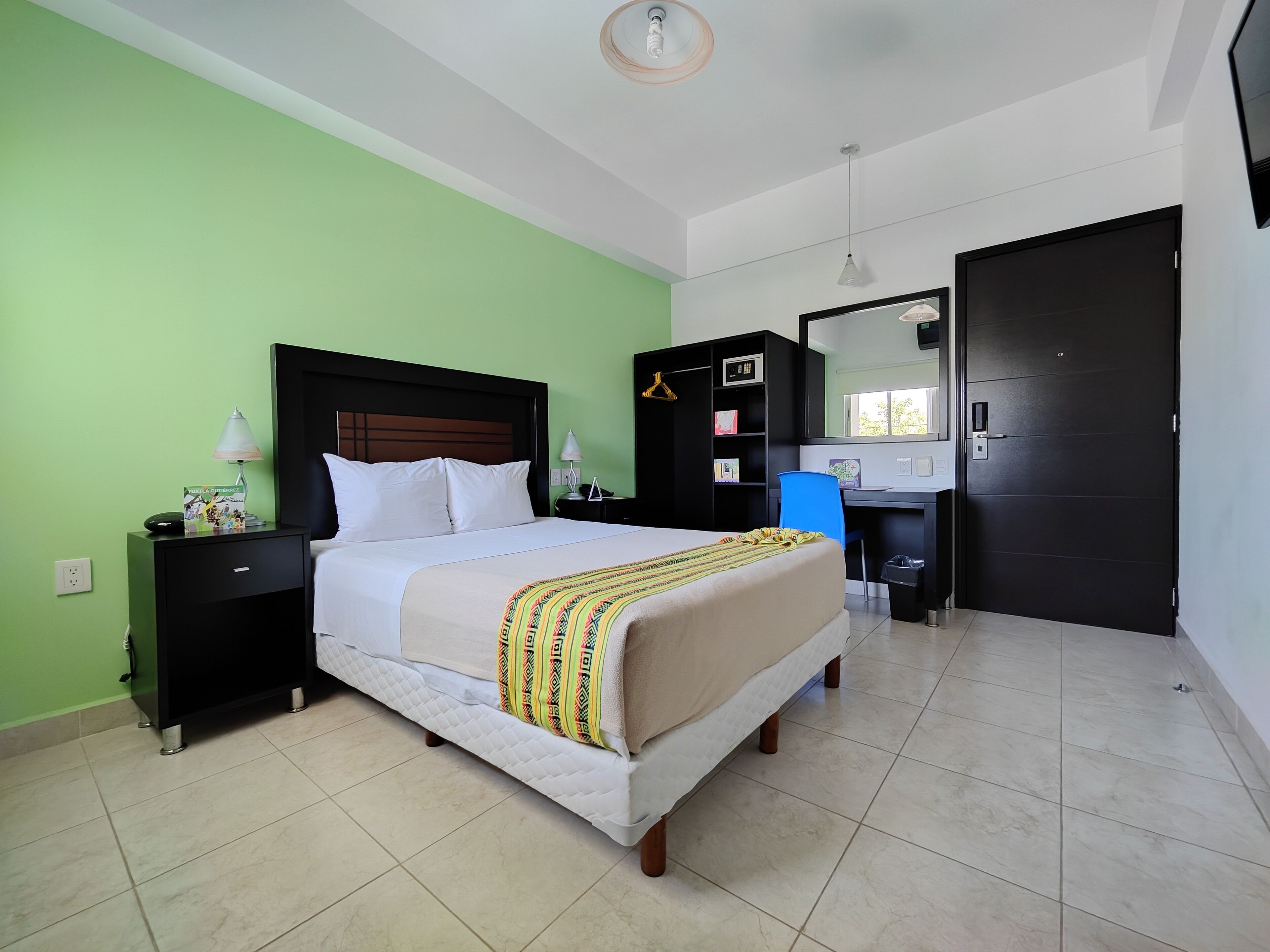 Quarto de hotel com cama, mesas de cabeceira, escrivaninha, cadeira, guarda-roupa e porta.