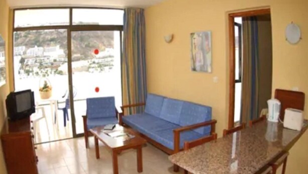 Una sala de estar con sofá, mesa de centro y mesa de comedor. Hay un televisor sobre un mueble bajo, y una ventana con cortinas.
