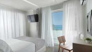 A modern hotel room with a bed, a chair, a TV, and a view of the sea. - Bella Mare (Rethymno)