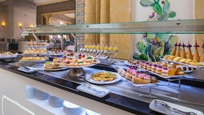 Un bufé con diversos alimentos, entre ellos pastelería, frutas y postres, exhibidos sobre un mostrador de vidrio con un mural decorativo en el fondo.