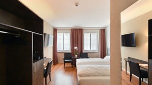 Ein Hotelzimmer mit Bett, Schreibtisch, Stuhl, Fernseher und einem Fenster mit Vorhängen.