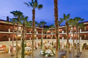 Un vestíbulo de hotel de varios pisos con palmeras, asientos al aire libre y una fuente central.