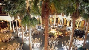 An indoor venue with multiple round tables and chairs, palm trees, and a central fountain. - Elba Palace Golf Boutique Hotel (Antigua)