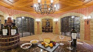 A wine tasting room with a brick wall, wooden ceiling, and a chandelier. There are wine bottles, cheese, and grapes on the table. - Elba Palace Golf Boutique Hotel (Antigua)