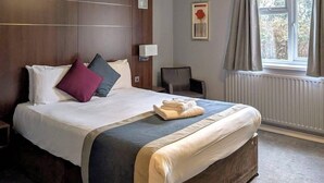 A hotel room with a large bed, a bedside table, a chair, a window with curtains, and a radiator. - Quality Hotel Coventry (Coventry)