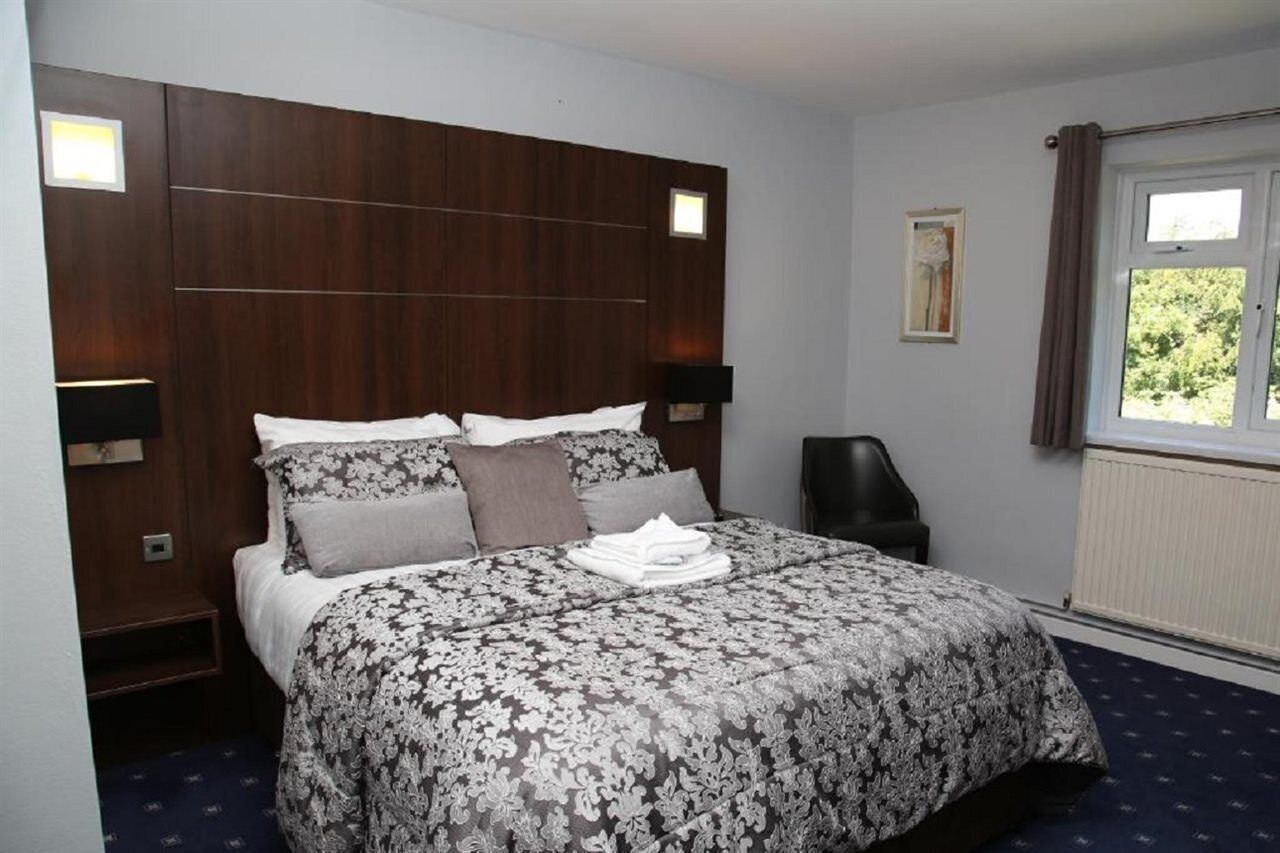 A hotel room with a large bed, dark wood headboard, two bedside lamps, a window with curtains, and a radiator.